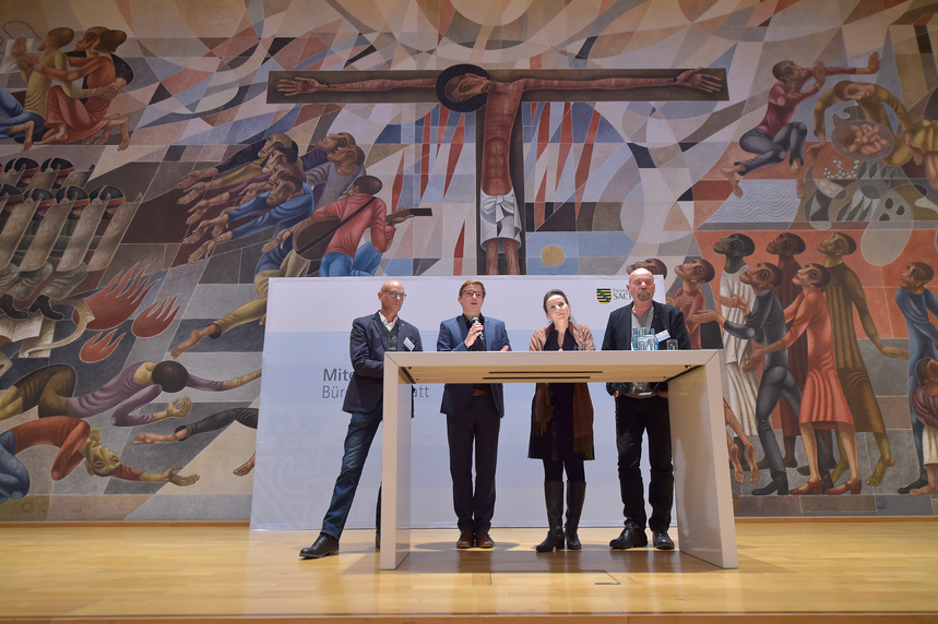 Drei Männer und eine Frau stehen an einem Tisch auf einer Bühne. Im Hintergrund befindet sich ein Wandgemälde.